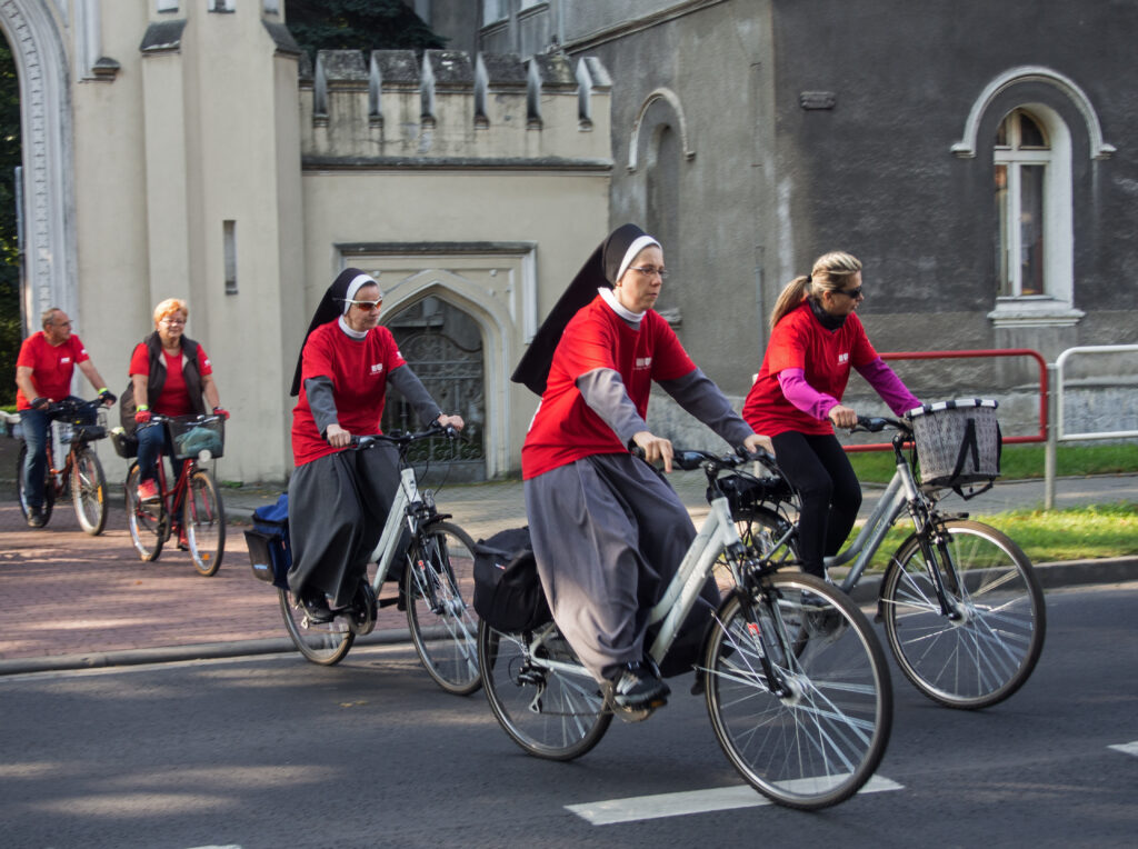 Siostra zakonna i kilka innych osób wyjeżdża na rowerach przez główną bramę pałacu.