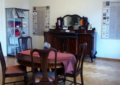 Na środku sali stół i drewniane krzesła. W tle drewniany, zabytkowy kredens z lustrem. Po prawej stronie plansza, po lewej gablota z orderami.