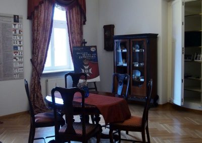 Na środku sali stół i drewniane krzesła, Z lewej strony okno z upiętymizasłonami i plakat z wystawy Chwała Rodu Henckel von Donnersmarck. Z prawej zabytkowa witryna z porcelana.