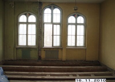 Trzy gotyckie okna. Wokół nich obdrapana ściana. Deski stropowe na podłodze.