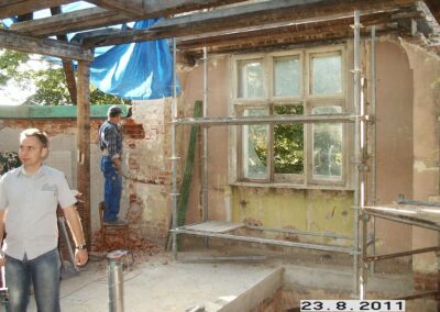 Mężczyźni w roboczych ubraniach remontujący walące się ściany. Przy ścianie z oknem stoi rusztowanie.