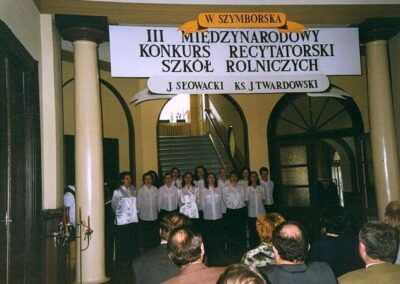 Grupa młodzieży w białych bluzkach stojących przy schodach. Nad nimi transparent z napisem W. Szymborska III Międzynarodowy Konkurs Recytatorski Szkół Rolniczych.