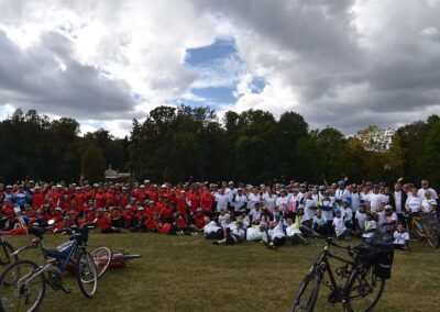Grupowe zdjęcie uczestników rajdu rowerowego. Z przodu po obu stronach stoją lub leżą rowery. Po lewej stronie grupa uczestników w czerwonych koszulkach, po prawej osoby w białych koszulkach. W tle drzewa i zachmurzone niebo.