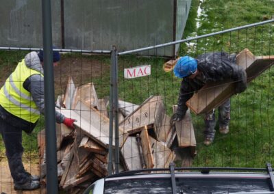 Mężczyzna w kamizelce odblaskowej i mężczyzna w kasku pochylają się nad materiałami budowlanymi przy ogrodzeniu.