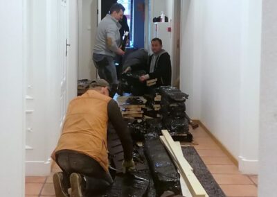 Czterech mężczyzn w wąskim korytarzu pochylonych nad materiałami budowlanymi.