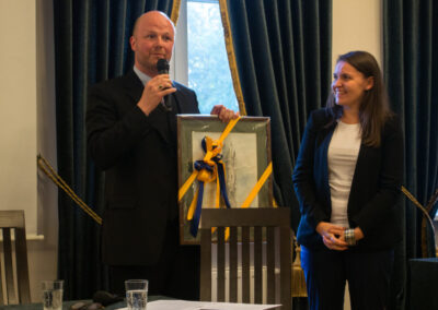 Po lewej stronie hrabia Mikołaj von Ballestrem mówi coś do mikrofonu trzymając w lewej ręce obraz w ramce przewiązany niebieską i żółtą wstążką. Po prawej stronie uśmiechnięta kobieta patrzy na hrabiego. Przed nimi stół z dwiema szklankami wody, kartką i długopisem.
