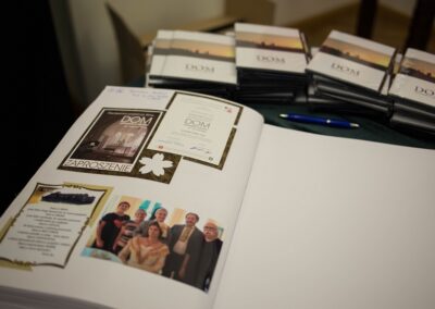 Z przodu otwarta księga, w której jest wklejone zdjęcie sześciu osób oraz zaproszenie na projekcję filmu "dom". Za księgą leży długopis i egzemplarze filmu.