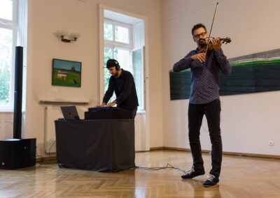 Z lewej mąarno gra na instumencie elektronicznym, po prawej mężczyzna grający na skrzypcach.żczyzna ubrany na cz