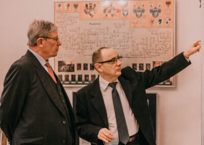 Dr Kuzio-Podrucki pokazuje hrabiemu eksponaty w gablocie, w tle plansza z drzewem genealogicznym.