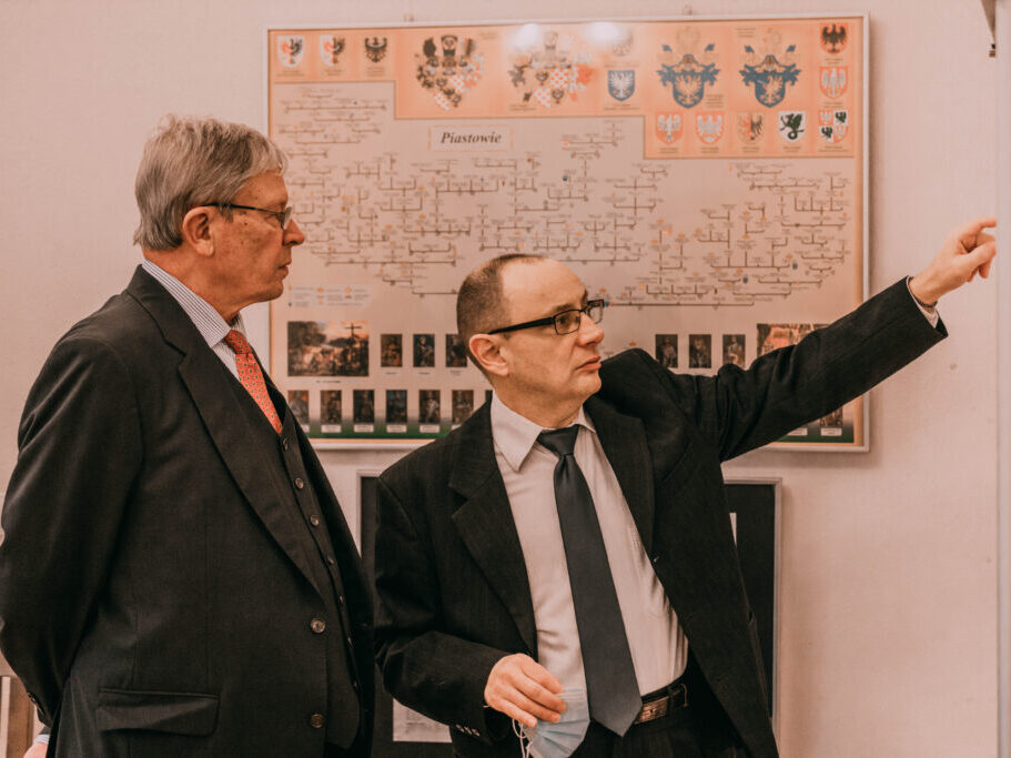Dr Kuzio-Podrucki pokazuje hrabiemu eksponaty w gablocie, w tle plansza z drzewem genealogicznym.