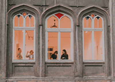 Fasada i okna pałacu z zewnątrz. W jednym oknie kobiety patrzące w obiektyw aparatu, jedna macha ręką. W drugim oknie kobiety rozmawiające ze sobą.
