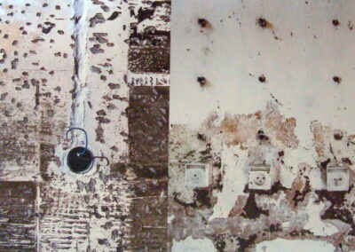 Obdrapana ściana z łuszczącą się farbą. Z lewej strony w scianie otwatra puszka elektryczna z wystającymi kablami. Po prawej na dole rząd gniazdek elektrycznych, nad nimi kikuty po uciętych, zardzewiałych drutach zbrojeniowych.