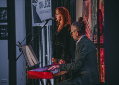 Adam Snopek gra na pianinie. Za nim Barbara Lubos śpiewa. W tle logo 100-lecie powstań slaskich.
