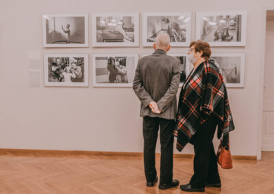 Starszy mężczyzna i starsza kobieta w kraciastej narzucie stoją przed powieszonymi na ścianie zdjęciami w antyramach.