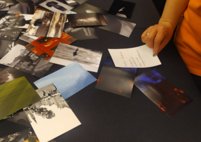 Na ciemnym stole leżą porozrzucane zdjęcia, z prawej strony widać rękę kobiety trzymającej kartkę z tekstem.
