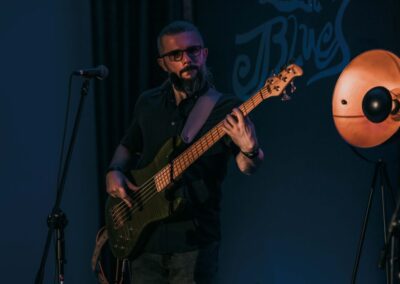 Za mikrofonem stoi Michał Rostański grający na gitarze basowej. Za nim biały napis "Blues".