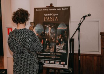 Kobieta we wzorzystej sukience stoi tyłem robiąc zdjęcie plakatu z wystawy Pasja.