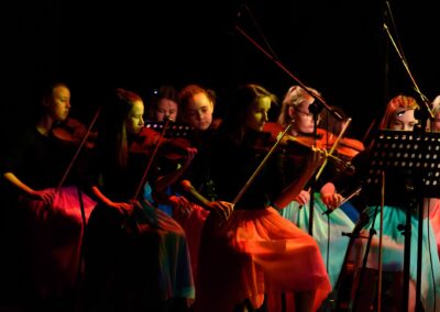 Grupa dziewczynek ubranych w czarne bluzki i jednokolorowe spódnice (czerwone, różowe, niebieskie) siedzących i grających w skupieniu na skrzypcach, Między nimi pulpity z nutami i stojaki mikrofonów