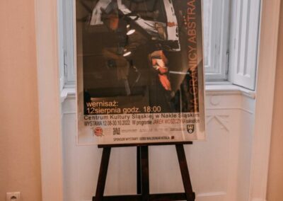 Na tle okna stoi na sztaludze plakat dotyczący wernisażu wystawy Rafała Stępniaka pod tytułem na granicy abstrakcji