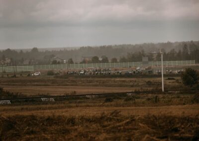 Zdjęcie tłumu uczestników jadących drogą przy polach, przez które biegną tory kolejowe. Zachmurzone niebo, mgła i padająca mżawka.