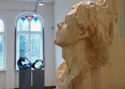 Głowa i twarz wyrzeźbiona w drewnie, w tle dwie rzeźby na postumentach i okno z zabytkowym witrażem.