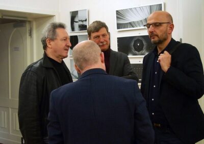 Czterech mężczyzn stoi i rozmawia, w tle widać czarno-białe zdjęcia wiszące na ścianie z wystawy "Siła detalu".