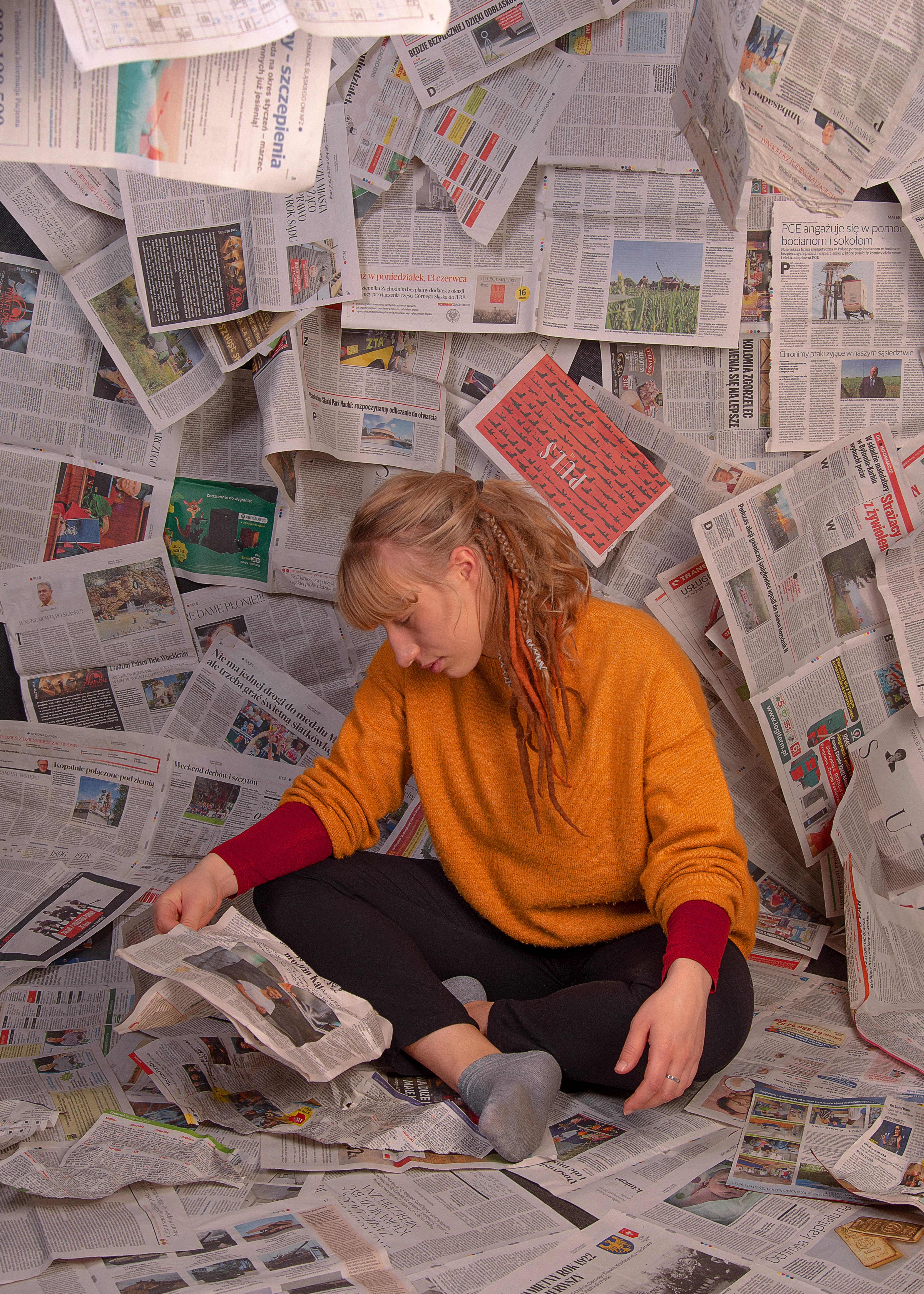 Kobieta siedząca na rozłozonych gazetach. Wokół niej na ścianach również wiszą gazety.