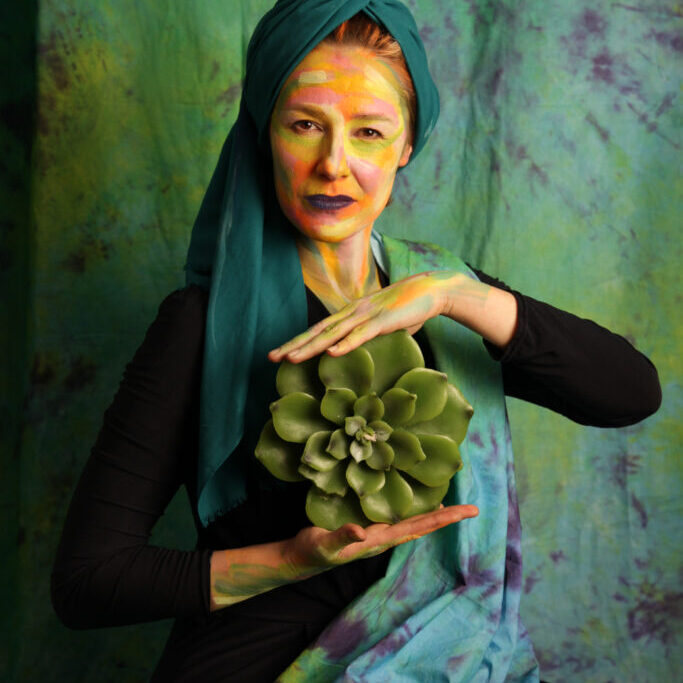 Kobieta w zielonym turbanie, z pomalowana na kolorowo twarzą, siedzi trzymając w dłoniach kwiat rozchodnika.