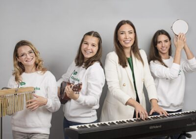 Cztery młode kobiety w białych bluzkach. jedna ma tamburyna, Druga stoi przy keyboardzie, trzecia gra na ukulele, cwarta na dzwonkach.