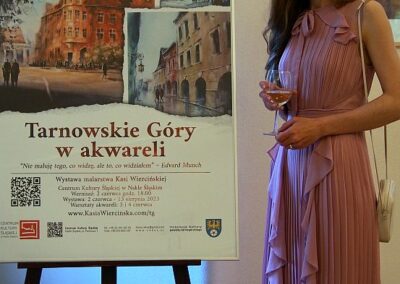 Kasia Wiercińska w długiej zwiewnej sukience, z kieliszkiem wina w ręce stoi obok plakatu z wystawy.