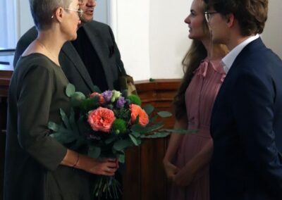 Mężczyzan i kobieta z bukietem kwiatów rozmawiają z autorka obrazów i stojacym obok niej mężczyzną.