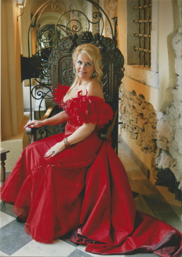 Gabriela de Silva w długiej czerwonej sukni z odsłonietymi ramionami, siesząca nakutym krześle.