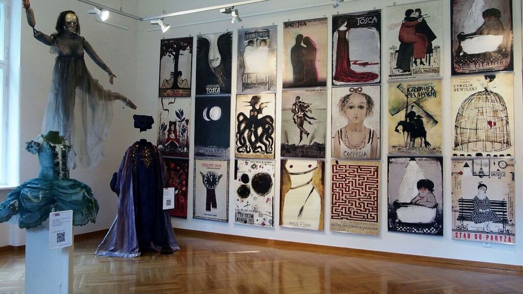 Trzy rzędy plakatów Ryszarda Kai do przedstawień operowych wiszące na białej ścianie. Z lewej podwisony manekin w zwiewnej sukni. Dwa stojące manekiny z kostumami i opisami.