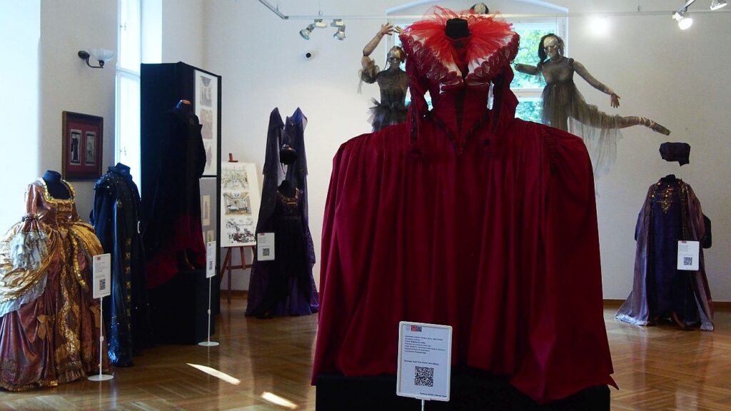 Manekin w  długiej czerwonej sukni z kryzą stojący na postumencie. W tle inne suknie i plakaty.