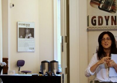 Anna Kempa-Gąsior stojaca obok otwartych drzwi, prowadzących do sali z cateringiem. Za na plakat Ryszarda Kai wiszący na ścianie.