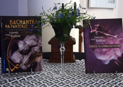 Stolik nakryty koronkową serwetą. Na środku drewniany młynek do kawy i wazonik z kwiatami. Po lewej stoi ksążka Gabrieli Kańtor Bachantka na Panterze, po prawej Tańcząc na czubkach palców.