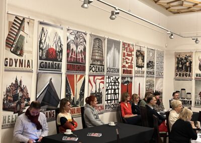 Ludzie siedzący przy stolikach nakrytych czarnym obrusem, pod ściana, na której wiszą plakaty Ryszarda Kai.