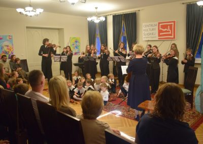 Krótkowłosa blondynka w granatowej sukni trzymająca skrzypce i stojąca przed grupą dzieci grających na skrzypcach.